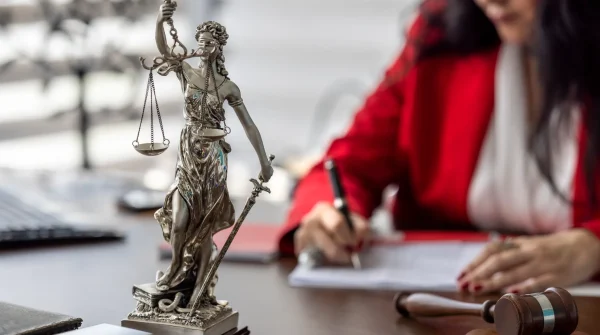 Justitia-Statue mit Waage und Schwert auf einem Schreibtisch, im Hintergrund eine Frau in rotem Blazer beim Unterschreiben juristischer Unterlagen – Symbol für Recht und Rechtsschutz.