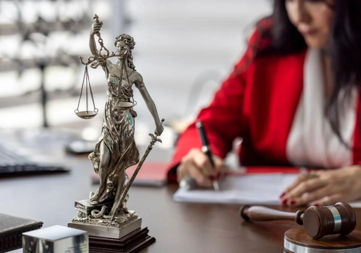 Justitia-Statue mit Waage und Schwert auf einem Schreibtisch, im Hintergrund eine Frau in rotem Blazer beim Unterschreiben juristischer Unterlagen – Symbol für Recht und Rechtsschutz.