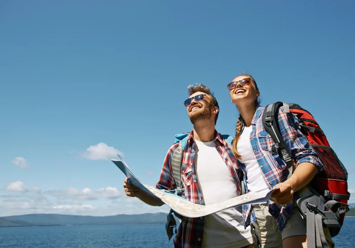 Junges Paar mit Wanderrucksäcken und Landkarte steht lachend am Wasser und plant seine Reise-und-Freizeit-Tour