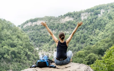 Frau sitzt mit ausgebreiteten Armen auf Felsen und blickt in bewaldete Berglandschaft während einer Wanderreise