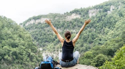 Frau sitzt mit ausgebreiteten Armen auf Felsen und blickt in bewaldete Berglandschaft während einer Wanderreise