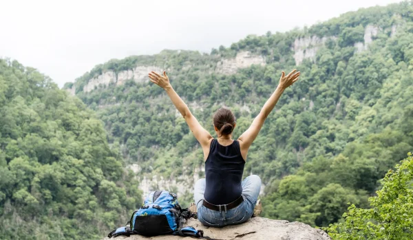 Frau sitzt mit ausgebreiteten Armen auf Felsen und blickt in bewaldete Berglandschaft während einer Wanderreise