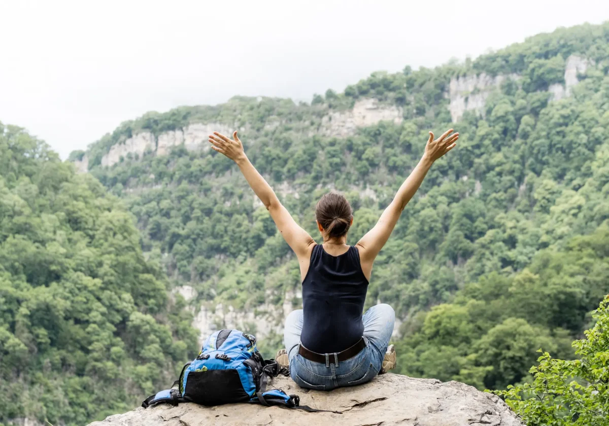 Frau sitzt mit ausgebreiteten Armen auf Felsen und blickt in bewaldete Berglandschaft während einer Wanderreise