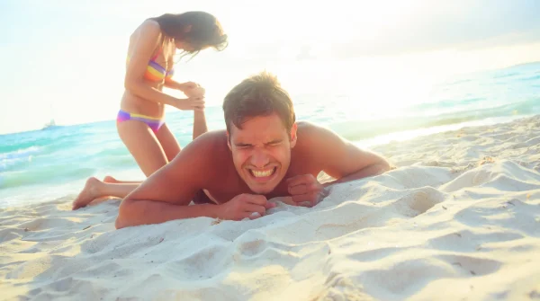 Mann mit schmerzverzerrtem Gesicht liegt verletzt im Sand, Frau hilft ihm am Strand – Thema Reiseunfallversicherung
