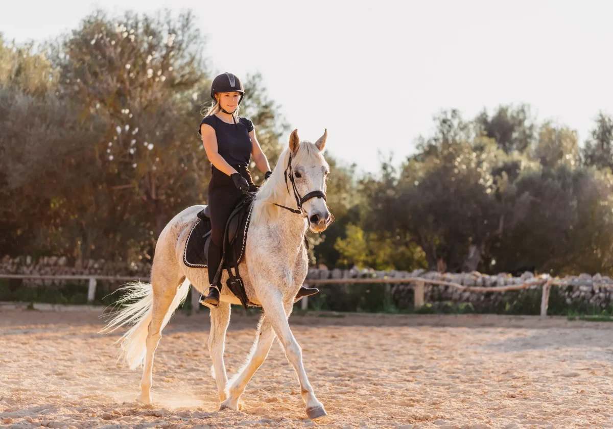 Reiterunfallversicherung Junge Reiterin mit Helm sitzt sicher auf einem weißen Pferd auf einem Reitplatz – Symbolbild für Unfallrisiken beim Reitsport.