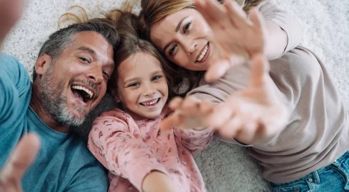 Lächelnde Familie mit Mutter, Vater und Tochter liegt auf dem Teppich und streckt lachend die Hände in Richtung Kamera – Symbol für Geborgenheit.