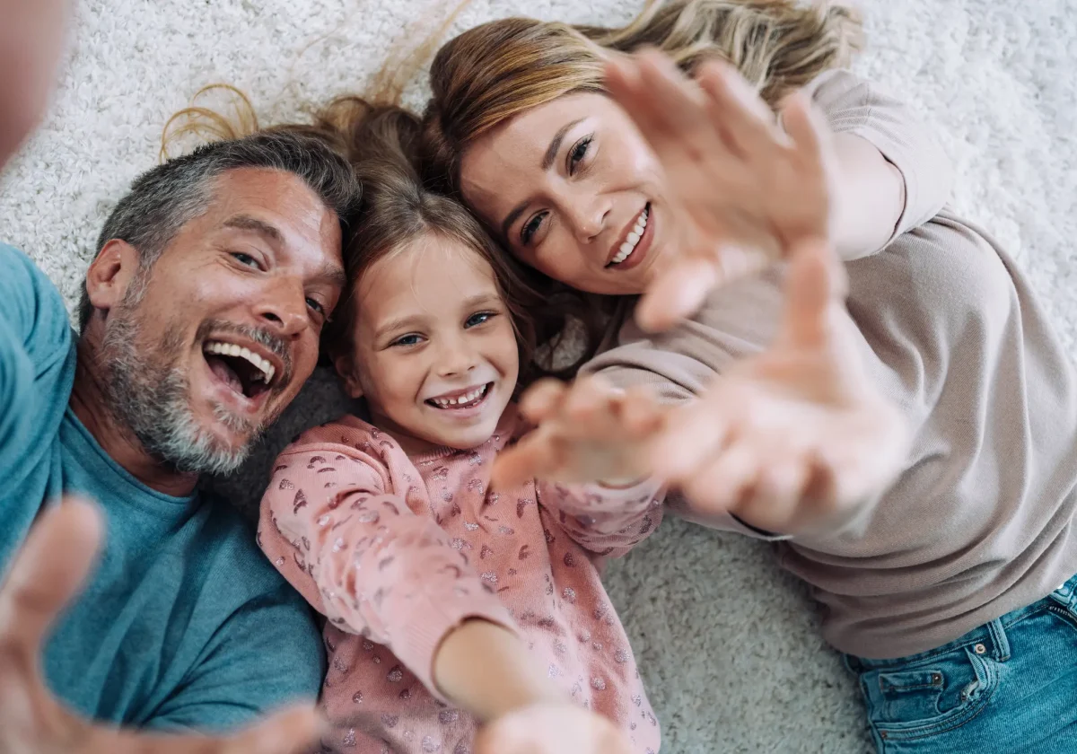 Lächelnde Familie mit Mutter, Vater und Tochter liegt auf dem Teppich und streckt lachend die Hände in Richtung Kamera – Symbol für Geborgenheit.