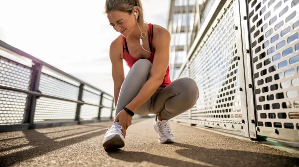 Junge Frau im Sportoutfit hält sich schmerzhaft den Knöchel nach einem Sturz beim Joggen – Sportunfall auf Brücke