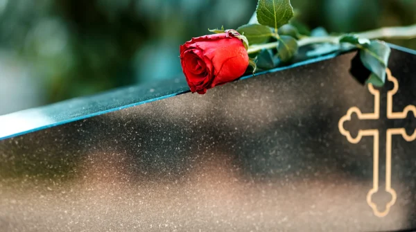 Rote Rose liegt auf einem dunklen Sarg mit goldener Kreuzverzierung, unscharfer Hintergrund, Symbol für Abschied und Erinnerung