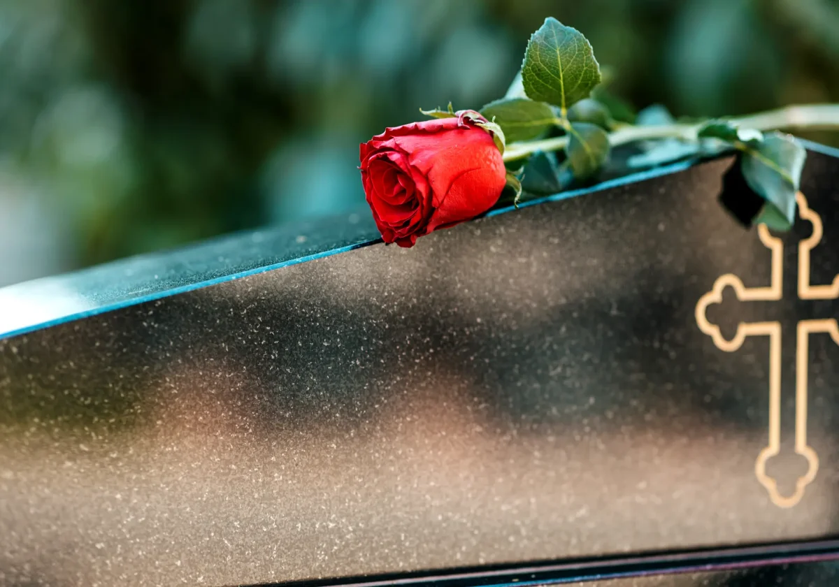 Rote Rose liegt auf einem dunklen Sarg mit goldener Kreuzverzierung, unscharfer Hintergrund, Symbol für Abschied und Erinnerung