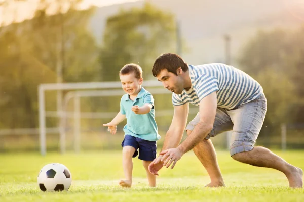 Vater spielt mit seinem kleinen Sohn auf dem Fußballfeld und schützt ihn mit einer privaten Unfallversicherung