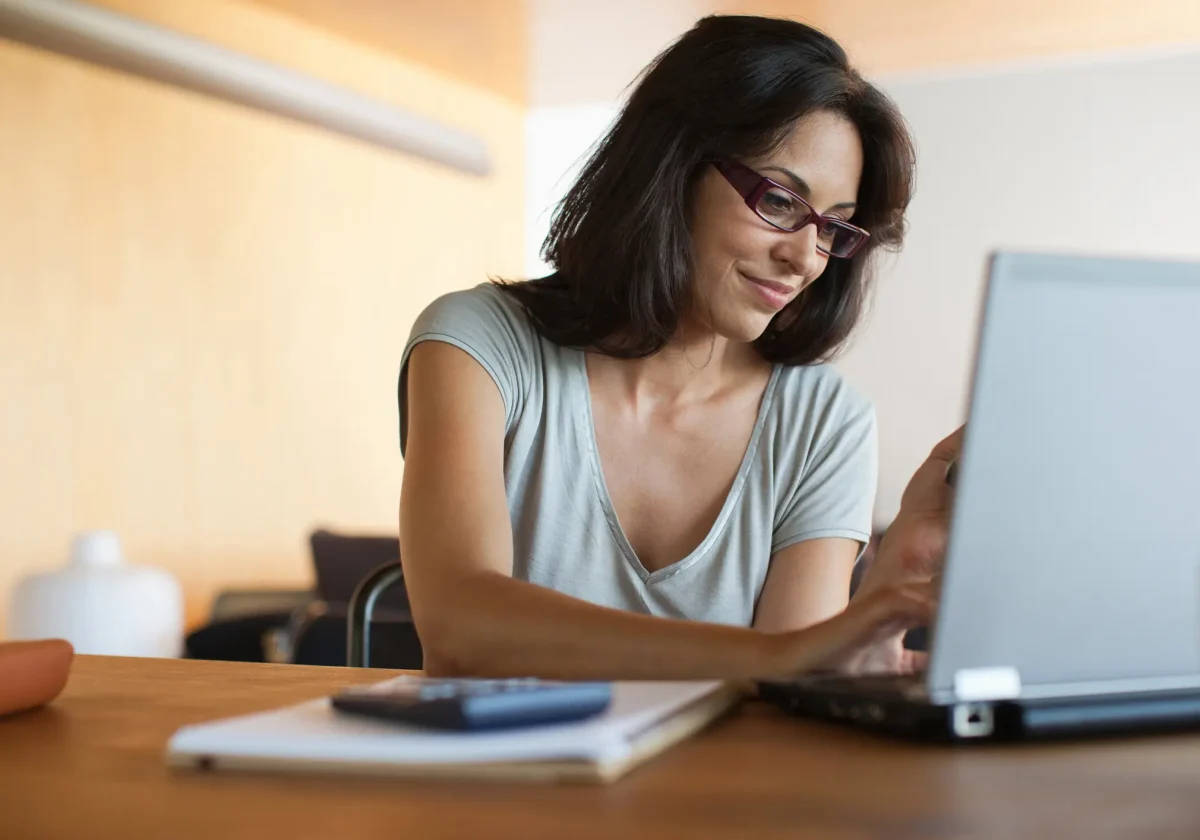 Frau mit Brille sitzt am Laptop und recherchiert, lächelt leicht – Notizbuch und Taschenrechner vor ihr