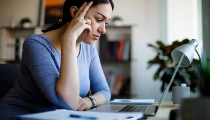 Junge Frau sitzt am Schreibtisch und blickt nachdenklich auf ihren Laptop, Hand an der Stirn, Unterlagen und Lampe im Vordergrund