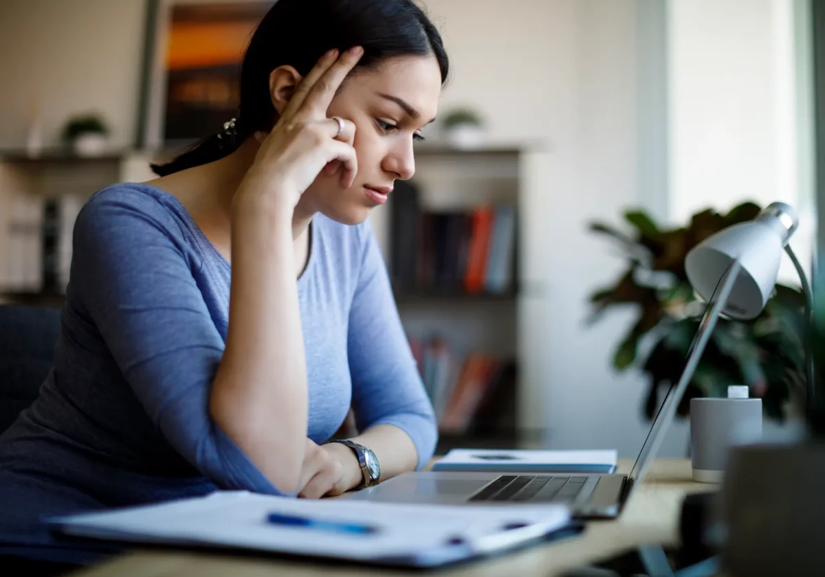 Junge Frau sitzt am Schreibtisch und blickt nachdenklich auf ihren Laptop, Hand an der Stirn, Unterlagen und Lampe im Vordergrund