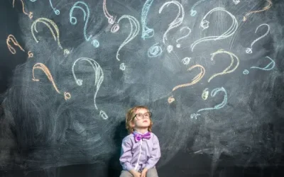 Kind sitzt vor Tafel mit vielen bunten Fragezeichen und blickt nachdenklich nach oben