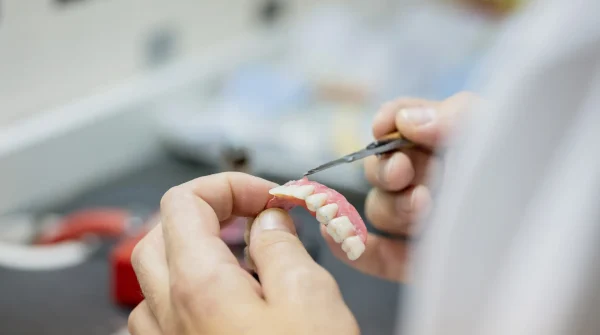 Zahntechniker hält Zahnprothese in der Hand und bearbeitet sie mit feinem Werkzeug, Fokus auf passgenauen Zahnersatz im Labor.