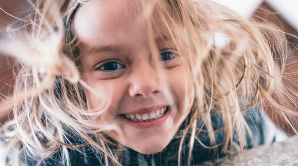 Lachendes Kind mit gesunden Zähnen und wehenden Haaren – Symbol für unbeschwertes Aufwachsen mit guter Zahnvorsorge