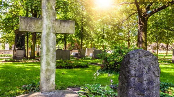 Grabkreuz und Grabstein auf einem sonnigen Friedhof mit viel Grün – symbolische Darstellung für eine anonyme Bestattung ohne Namensnennung