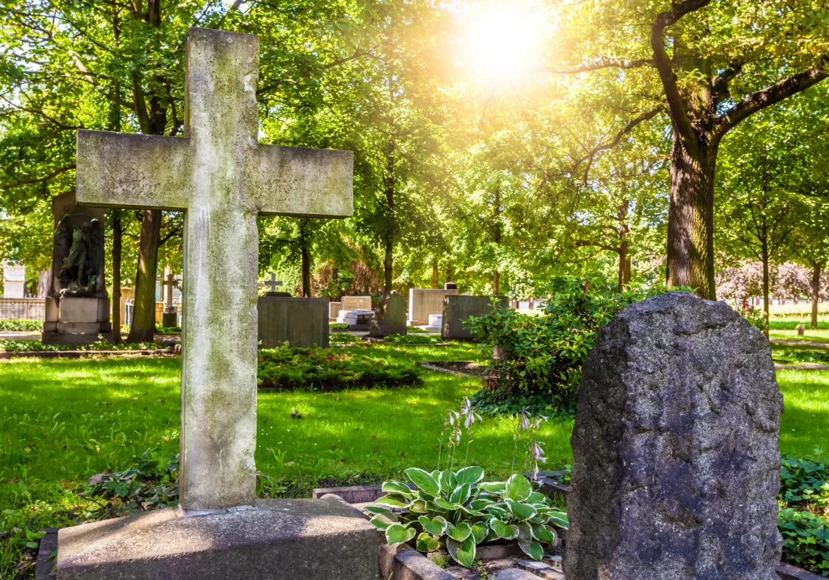 Grabkreuz und Grabstein auf einem sonnigen Friedhof mit viel Grün – symbolische Darstellung für eine anonyme Bestattung ohne Namensnennung