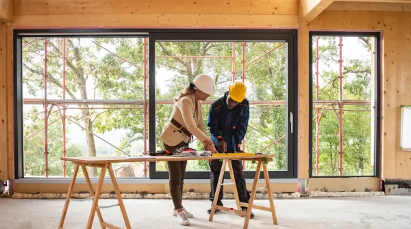 Zwei Bauleiter mit Schutzhelm besprechen Pläne auf einer Baustelle mit großem Fenster und Holzwänden – Sicherheit und Baukoordination