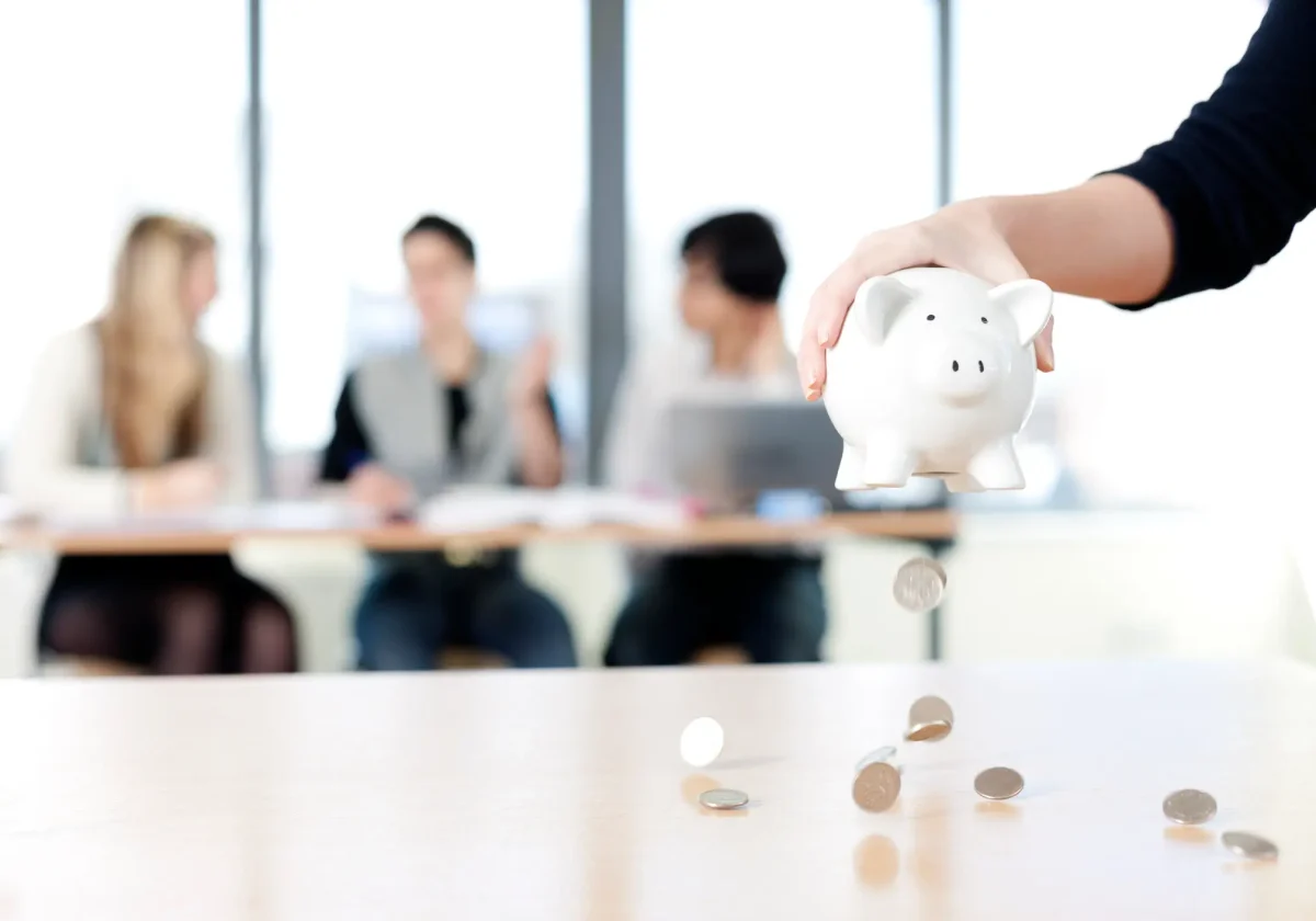 Hand hält ein Sparschwein über einem Tisch, Münzen fallen herunter – im Hintergrund sitzen Menschen im Büro