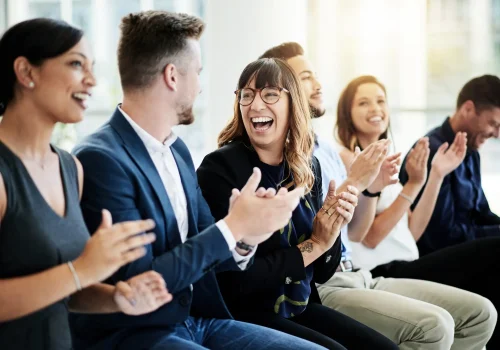 Junge Berufstätige sitzen nebeneinander im Büro, lachen gemeinsam und applaudieren in einer entspannten Teamatmosphäre