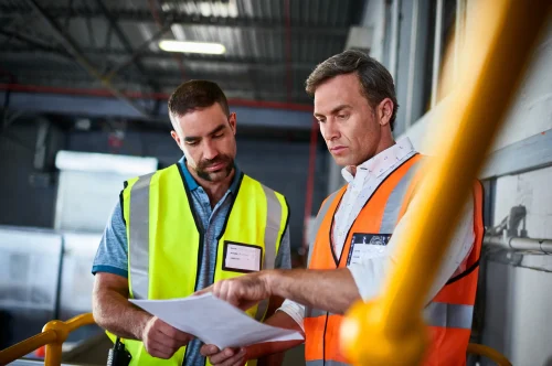Zwei Männer in Warnwesten besprechen Unterlagen in einem Industriebetrieb