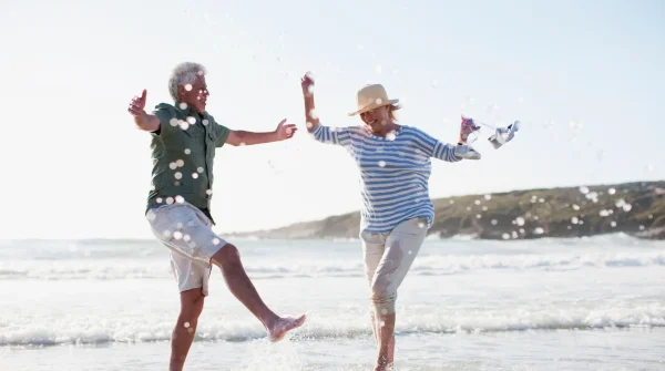 Älteres Paar läuft lachend durchs Wasser am Strand – Symbolbild für früher in Rente und erfüllten Ruhestand
