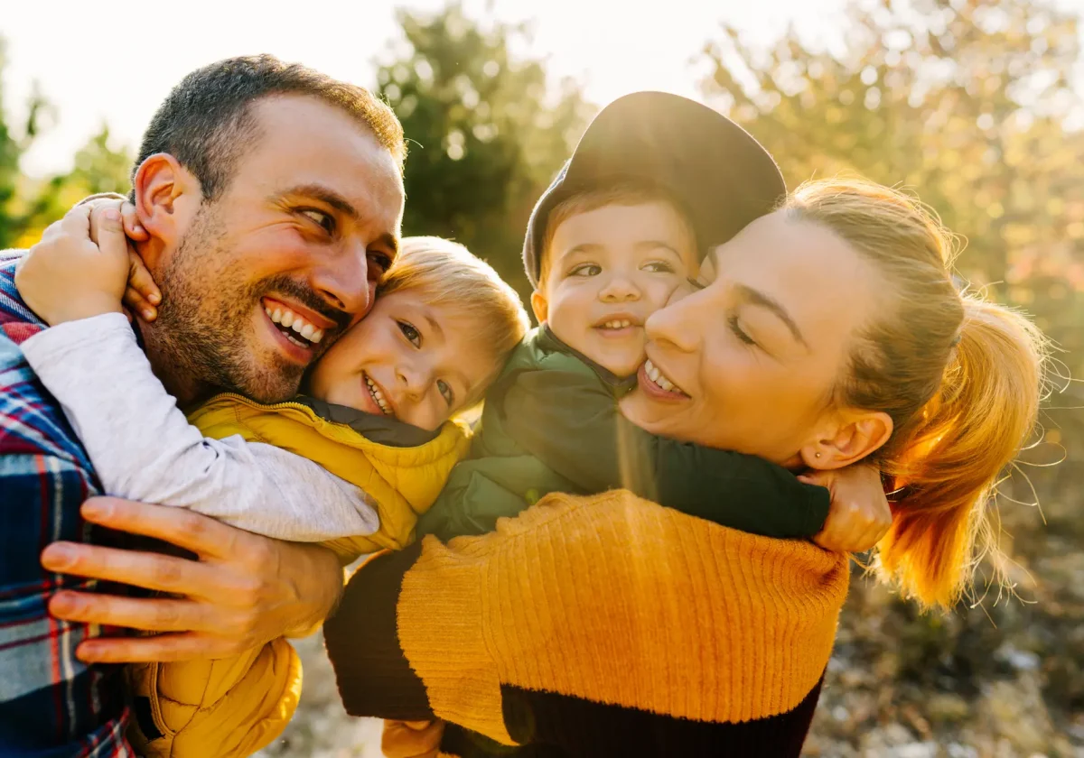 Gesundheit-Pflege Glückliche Familie mit zwei kleinen Kindern umarmt sich lachend im Sonnenlicht im Freien