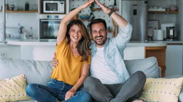 Lachendes Paar sitzt auf dem Sofa und formt mit den Händen ein Hausdach – Symbol für Sicherheit rund um Haus und Wohnen