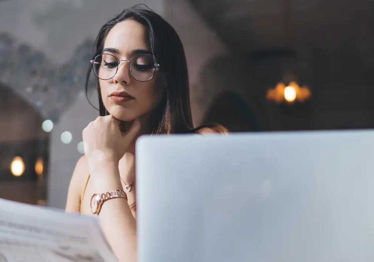 Junge Frau mit Brille liest aufmerksam eine Zeitung in einem Café, vor ihr steht ein Laptop, im Hintergrund warme Lichtquellen