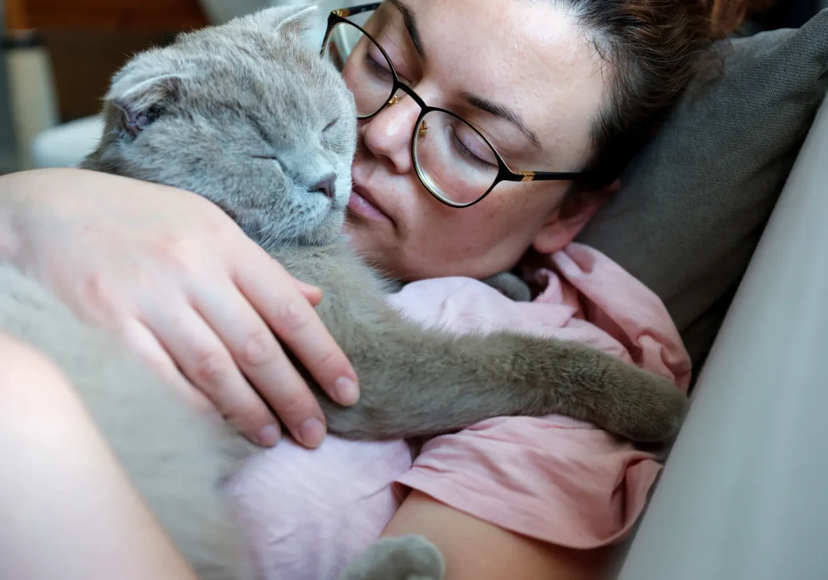 Katzenkrankheiten Eine Frau hält ihre kranke Katze liebevoll im Arm – beide liegen auf dem Sofa, eng aneinandergeschmiegt und in einem ruhigen Moment vereint.