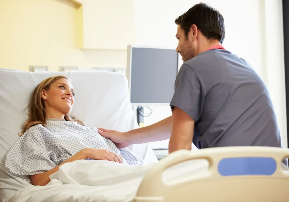 Krankenhauszusatzversicherung Lächelnde Patientin liegt im Krankenhausbett und spricht mit einem Arzt in moderner Klinikumgebung