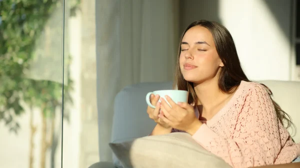 Junge Frau sitzt entspannt auf dem Sofa, genießt mit geschlossenen Augen eine Tasse Tee im Sonnenlicht