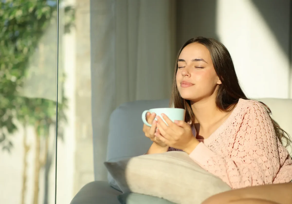 Junge Frau sitzt entspannt auf dem Sofa, genießt mit geschlossenen Augen eine Tasse Tee im Sonnenlicht