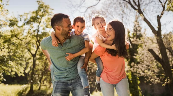 Lachende Familie mit zwei Kindern auf dem Rücken der Eltern bei einem Spaziergang im Grünen an einem sonnigen Tag