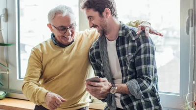 Junger Mann zeigt älterem Vater etwas am Smartphone, beide lachen und wirken vertraut und gut gelaunt