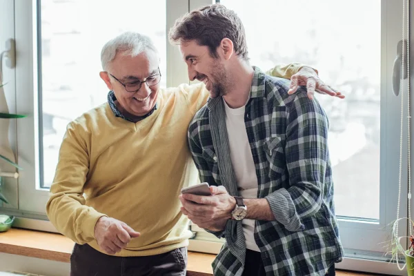 Junger Mann zeigt älterem Vater etwas am Smartphone, beide lachen und wirken vertraut und gut gelaunt