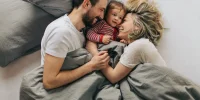 Junge Familie liegt gemeinsam im Bett und lacht, Mutter, Vater und Baby blicken sich liebevoll an