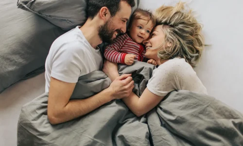 Junge Familie liegt gemeinsam im Bett und lacht, Mutter, Vater und Baby blicken sich liebevoll an