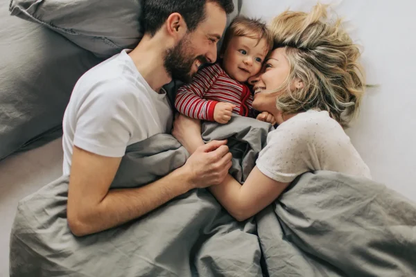Junge Familie liegt gemeinsam im Bett und lacht, Mutter, Vater und Baby blicken sich liebevoll an