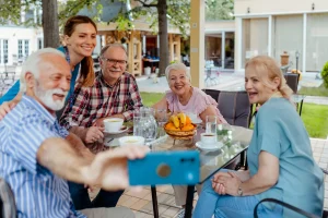 Seniorengruppe sitzt lachend an einem Gartentisch, eine Pflegekraft macht ein gemeinsames Selfie mit dem Smartphone