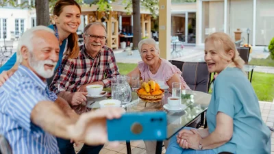 Seniorengruppe sitzt lachend an einem Gartentisch, eine Pflegekraft macht ein gemeinsames Selfie mit dem Smartphone