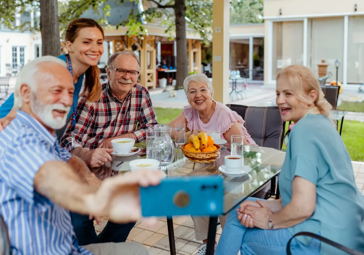 Pflegezusatzversicherung Seniorengruppe sitzt lachend an einem Gartentisch, eine Pflegekraft macht ein gemeinsames Selfie mit dem Smartphone