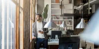Selbstständiger Mann steht in modernem Büro am Fenster und blickt nachdenklich hinaus, Tablet in der Hand