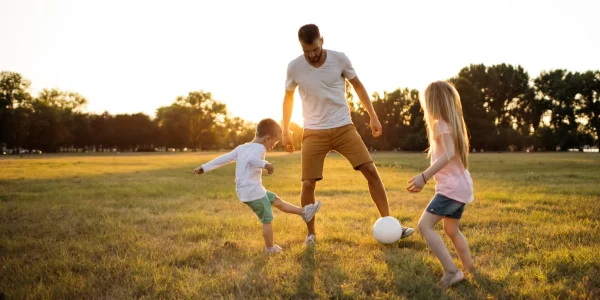 Vater spielt bei Sonnenuntergang mit seinen zwei Kindern Fußball auf einer Wiese
