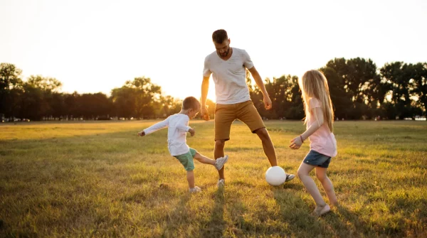 Vater spielt bei Sonnenuntergang mit seinen zwei Kindern Fußball auf einer Wiese