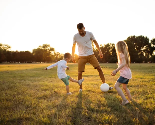 Vater spielt bei Sonnenuntergang mit seinen zwei Kindern Fußball auf einer Wiese
