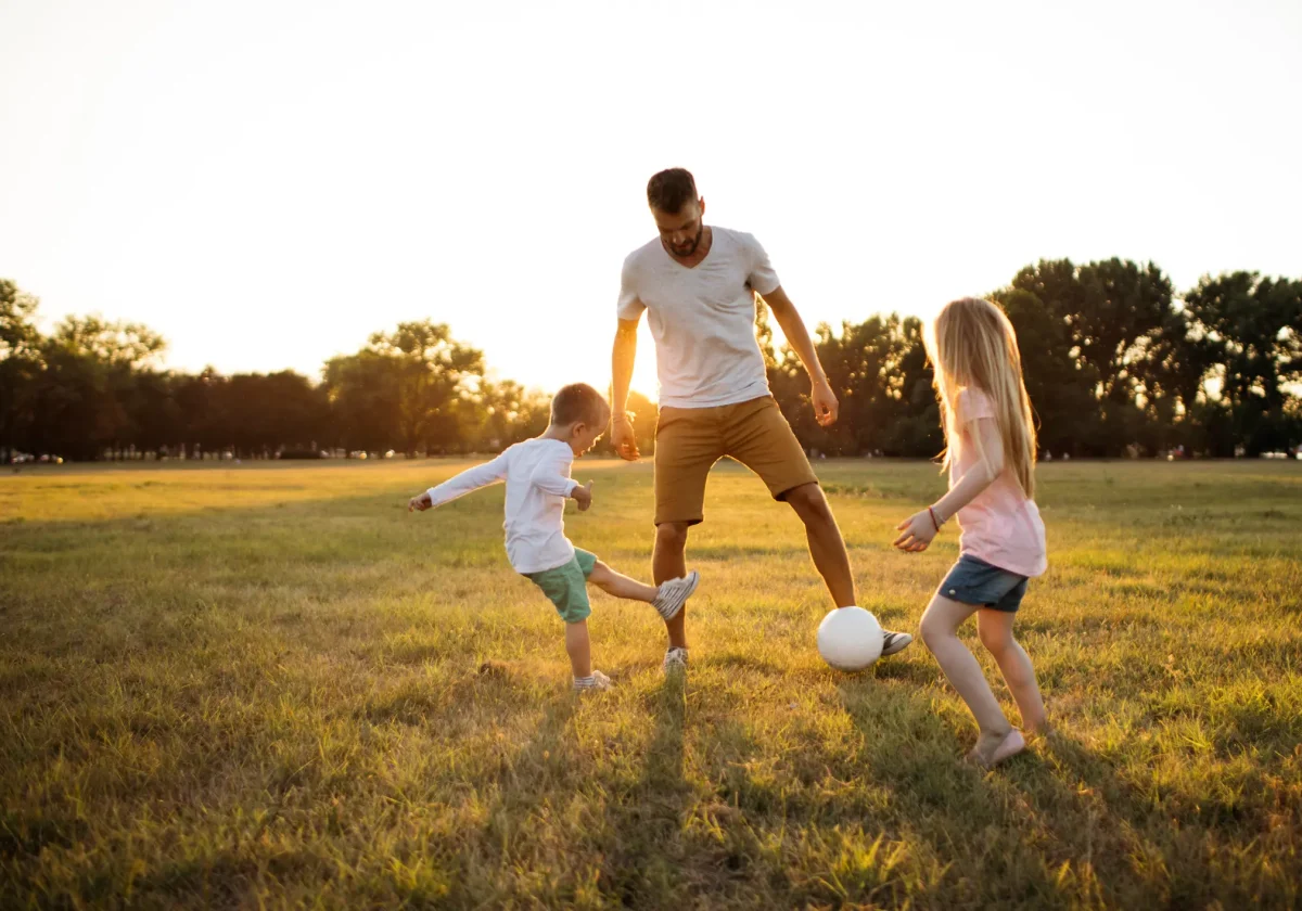Vater spielt bei Sonnenuntergang mit seinen zwei Kindern Fußball auf einer Wiese