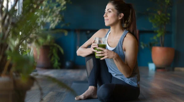 Frau sitzt entspannt auf einer Yogamatte und hält einen grünen Smoothie – Symbol für Gesundheit und private Vorsorge